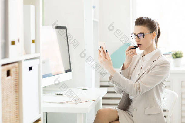 <strong>年轻</strong>的女商人使用她的智能手机作为镜子, 同时应用口红的工作场所