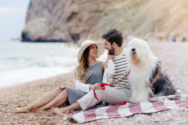 年轻夫妇在爱休息与毛茸茸的狗坐在沙滩上的格子上, 微笑着看着对方