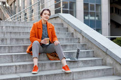 在城市场景中穿着鲜艳橙色外套的时髦女士，使用笔记本电脑，在休息的时候在电脑上工作。室外技术。喝咖啡、茶、可可