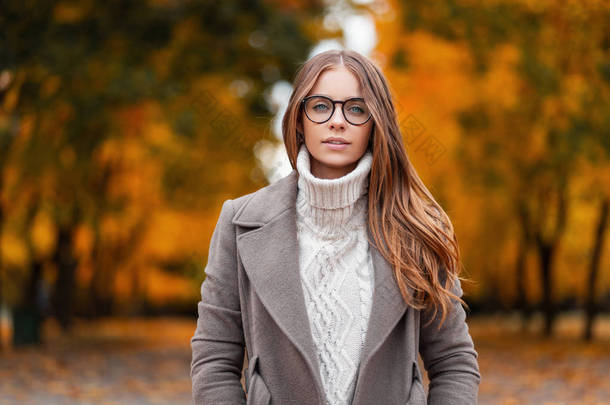 一个戴着时髦眼镜、穿着针织时尚白色毛衣、穿着雅致外套的漂亮年轻女子在公园里的秋天画像，她的背景是树叶呈橙色的树木