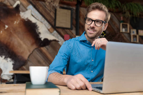 有魅力的商人，戴着<strong>眼镜</strong>，坐在办公桌前，在笔记本电脑上工作，一边高兴地看着远处的咖啡店