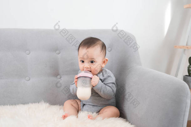 可爱的小女孩坐在沙发上, 从瓶子里喝着牛奶, 微笑着。快乐的婴儿。家庭人室内的内部概念。童年的最佳时间!