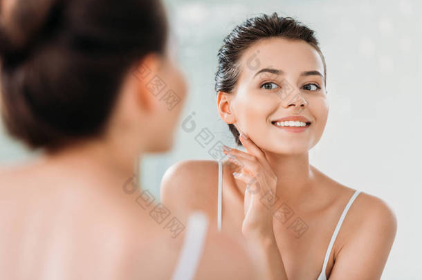 <strong>美丽</strong>的微笑的年轻妇女触摸皮肤, 看着镜子在浴室里