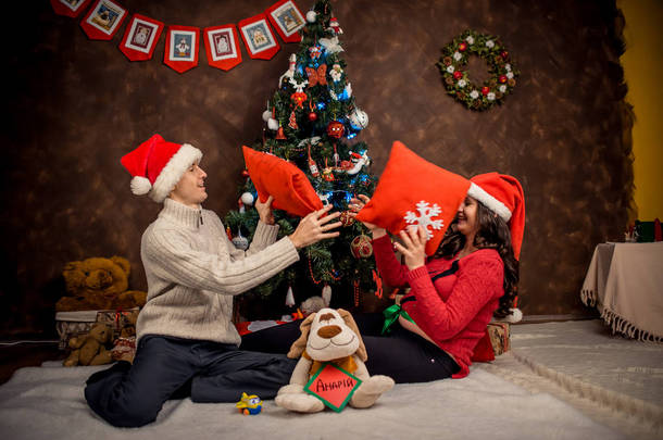 快乐的室内家庭人像圣诞老人的帽子。美丽的孕妇正在与她的丈夫在装饰圣诞树附近玩枕头.