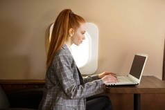 美丽的年轻女孩与红头发是在商务舱飞机与笔记本电脑飞行