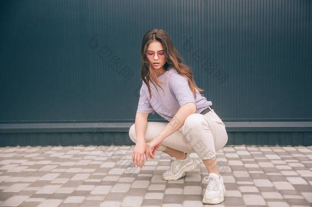 年轻时髦的女人被灰色的蓝色背景隔离了。迷人的女孩穿着紫色衬衫和白色裤子，蹲着坐着，看着镜头。流行的立场.