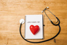 听诊器顶视图，笔记本上装饰心脏，世界健康日字母木制背景