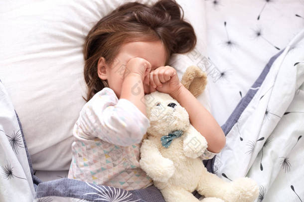 顶视图的小女孩躺在床上与泰迪熊，<strong>心情</strong>不好，不想上去和亲切的花环，蹒跚学步的孩子在枕头上揉她的眼睛，看起来很伤心。童年概念.