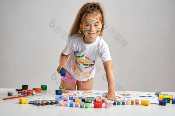 穿着白色T恤的小女孩站在桌旁，手里拿着什么东西和彩绘，手绘在上面。被白色隔离了中特写镜头.