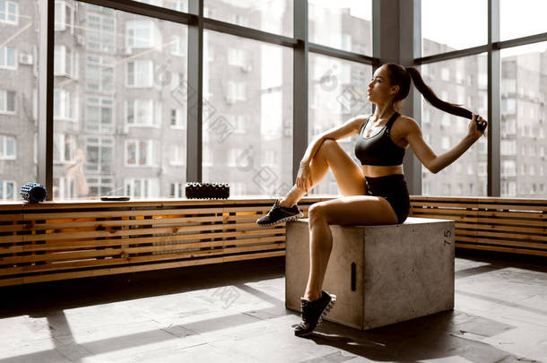 美丽的苗条女孩穿着黑色运动上衣和短裤坐在一个木箱在阳光下在健身房的窗口前