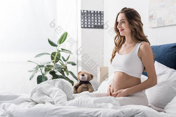 很高兴怀孕的年轻女子<strong>坐在床上</strong>摸着肚子