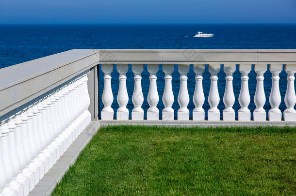 带绿色草坪和石栏杆的露台，背景是蓝天，水上有大海和游艇，是生态友好型建筑的细节.