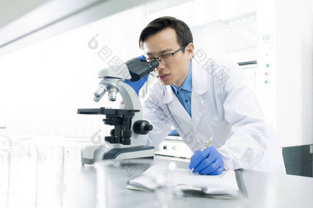 身穿白大褂的亚洲医学家用显微镜检查标本，水平肖像