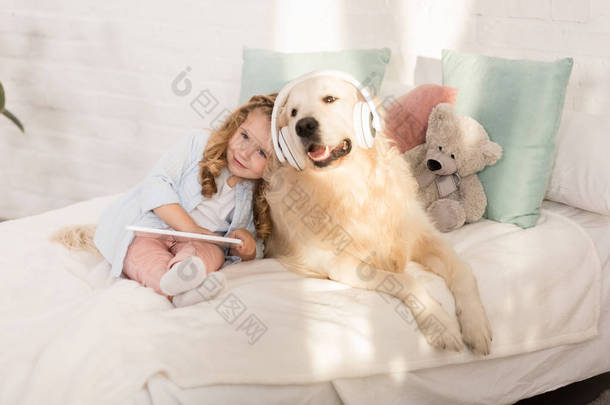 可爱的孩子拿着平板电脑, 靠在金毛猎狗身上, 耳机躺在儿童房的床上