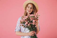 一个快乐的年轻女子在夏季礼服和草帽持有康乃馨花束隔离粉红色背景的肖像