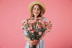 一个快乐的年轻女子在夏季礼服和草帽给康乃馨花束隔离粉红色背景的肖像