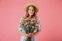 一个可爱的年轻女子在夏季礼服和草帽持有康乃馨花束隔离粉红色背景的肖像
