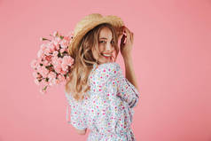 在夏季礼服和草帽捧着康乃馨花束, 看着她的肩膀被孤立的粉红色背景的一个迷人的年轻女子的肖像