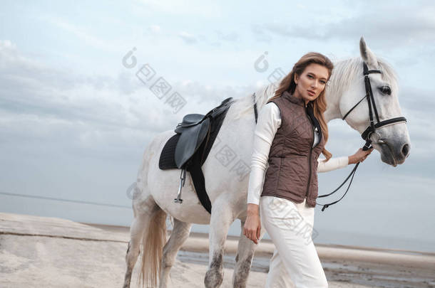 穿着时尚骑师服装的迷人而美丽的年轻女子,在海边靠近水面时,拿着马,摆出<strong>姿势</strong>。一个有吸引力的骑<strong>手</strong>在外面摆<strong>姿势</strong>。自然景观