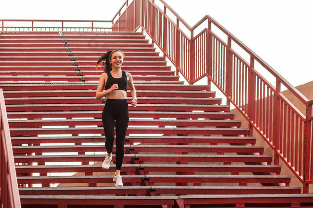 跑步运动员，穿着黑色运动服，紧身裤和上衣，适合快乐的女孩，跑下楼梯进行有氧训练，慢跑锻炼减肥。保健概念、户外运动