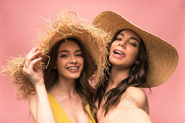 两个微笑的女孩在草帽采取自拍孤立在粉红色
