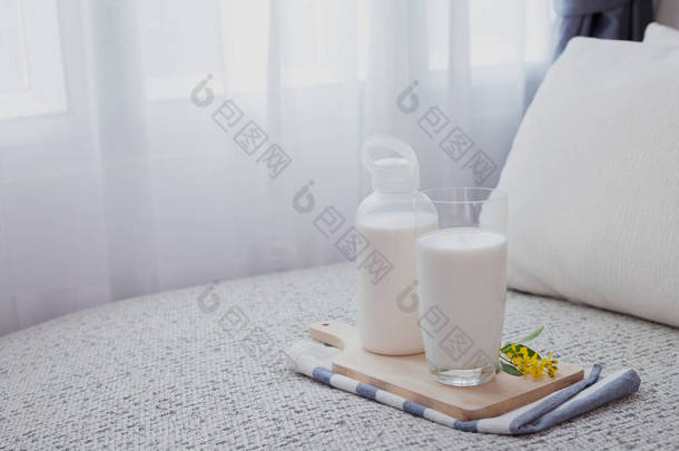 一瓶牛奶和一杯牛奶放在带白色窗帘窗的客厅的床上。正面视图.