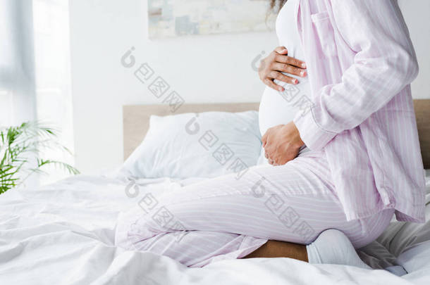 裁剪视图怀孕的非洲美国妇女<strong>坐在床上</strong>和触摸肚皮 