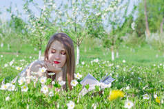 年轻漂亮的女人在白色的顶部长头发躺在草地上的花朵在苹果树花园和阅读书在阳光明媚的一天。复制文本的空间