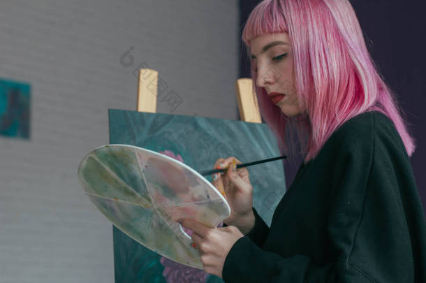 华丽的女艺术家与粉红色的头发工作的艺术项目与调色板在她的手中的艺术工作室。年轻女子画一幅画