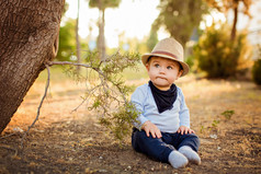 一顶草帽和蓝色裤子坐在一起的可爱的小宝贝男孩撅起嘴唇，夏季日落在地球上的一棵树旁