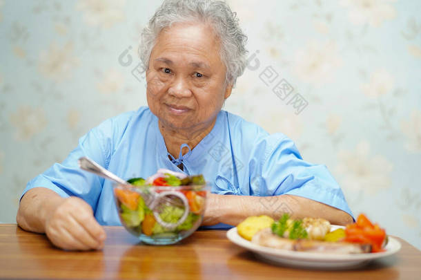 亚洲老人或老年老太太妇女病人吃早餐健康的食物与希望和快乐, 而坐在病床上, 饿在医院