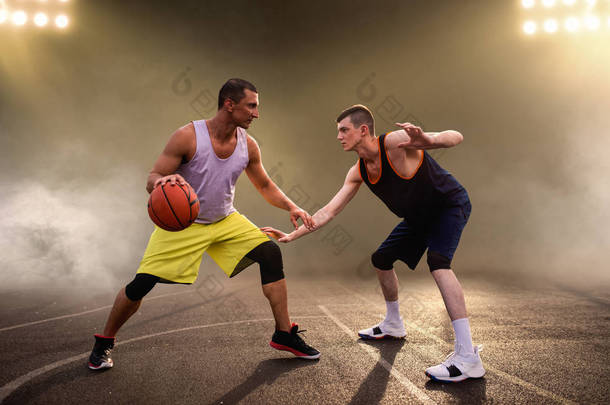 两名篮球运动员在球场上<strong>打球</strong>，背景漆黑，灯光明亮。 男子运动员穿着运动服在街头篮球训练中比赛