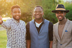 在夏天的公园里，一群穿着时髦西装的三个黑人男子。 非洲裔美国人的朋友们，他的恐慌商人在户外打电话给自己拍照