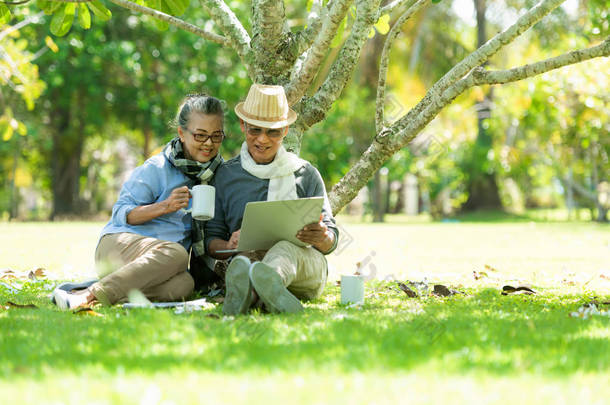 亚洲生活方式老年夫妇喝咖啡和网上购物在自然公园快乐和放松的时间。老年家庭休息和退休后的寒意.