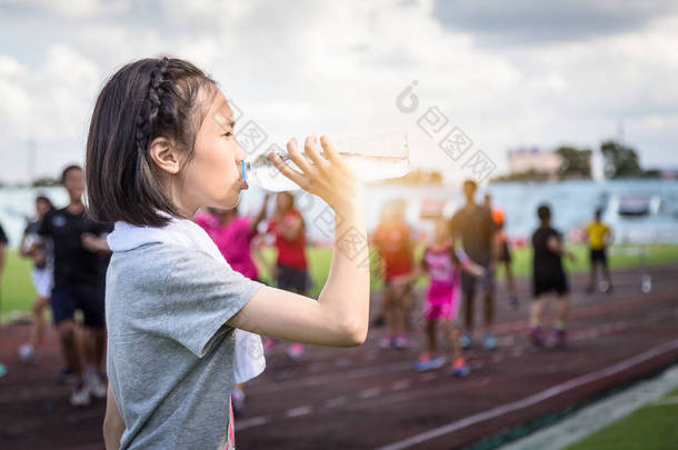 亚洲女孩拿着一瓶水，在阳光下从<strong>塑料瓶</strong>喝水的肖像，女运动员在跑步后休息，喝水，以弥补汗水损失，减少在炎热的晴天口渴 