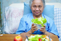 亚洲老人或老年老太太妇女病人吃早餐健康的食物与希望和快乐, 而坐在病床上, 饿在医院