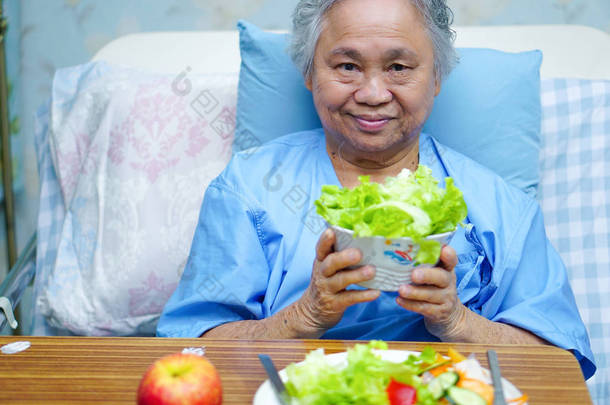 亚洲老人或老年老太太妇女病人吃早餐健康的食物与希望和快乐, 而坐在病床上, 饿在医院