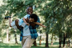 微笑的非洲裔美国人父亲抱着n臂可爱的儿子在绿色公园 