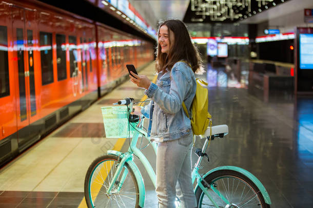 背着背包和自行车的少女站在<strong>地铁站</strong>，手里拿着智能手机，滚动着发短信，微笑着笑。未来明亮的<strong>地铁站</strong>。芬兰， 埃斯波
