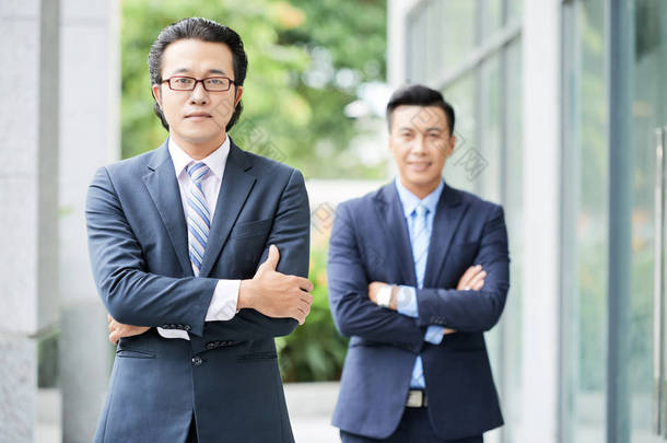 亚洲两个自信的商人穿着西装站在<strong>双臂</strong>交叉，看着相机户外的肖像