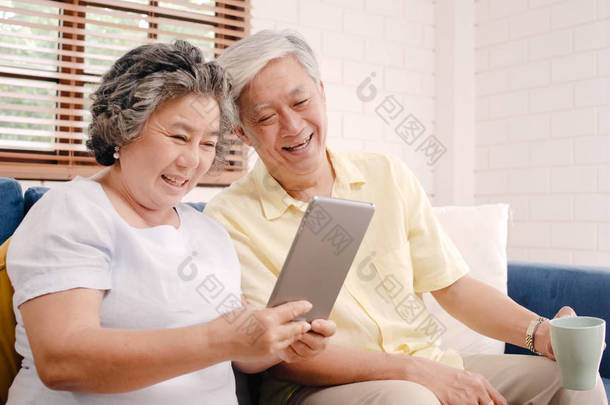 亚洲老年夫妇在家里的客厅里使用平板电脑和喝咖啡, 在家里放松的时候躺在沙发上享受爱情时刻。享受时间生活方式的老年家庭在家里的概念.
