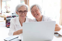幸福的两个亚洲老妇人，姐妹或朋友在家里交谈和享受使用笔记本电脑在一起，微笑的老人和她的友谊互相牵手，技术和朋友的概念