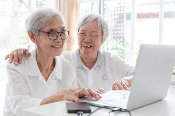 幸福的两个亚洲老妇人，姐妹或朋友在家里交谈和享受使用笔记本电脑在一起，微笑的老人和她的友谊互相牵手，技术和朋友的概念