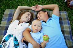 幸福的家庭：妈妈、爸爸和宝宝躺在绿草上的毯子上，手牵手。一个手里拿着苹果的孩子抬起来，父母互相看着对方。顶部的视图。概念