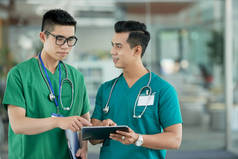 两名身穿医疗擦边球的亚洲年轻男子站在医院大厅模糊的背景上, 指着平板电脑上的信息
