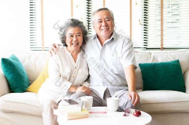 一对快乐的亚洲老年夫妇在家里的沙发上放松的肖像, 妻子抱着丈夫对着镜头微笑