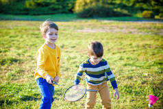 两个活跃的学龄前男孩夏天在室外场地打羽毛球。孩子们打网球。儿童学校体育活动。儿童运动员的网球拍和羽毛球运动。朋友们在一块温暖的田野里快乐.