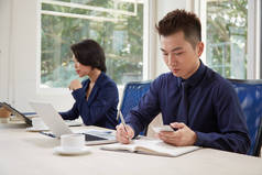 认真的年轻商人坐在桌旁, 在他的规划师的工作中, 用智能手机做笔记, 他的同事在他身边工作