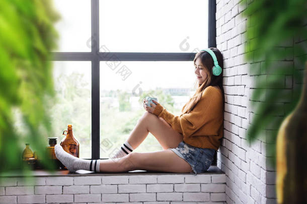 年轻的女人放松与音乐从耳机在舒适的房子附近的窗口, 享受早晨阳光和热咖啡