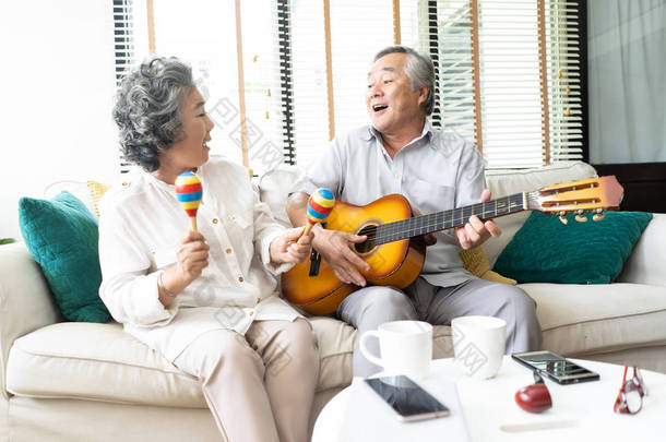 客厅里的情人。微笑的老人弹吉他和她的妻子拿着马拉卡斯跳舞和坐在家里的沙发有趣的肖像, 活动家庭的爱和生活风格的概念.